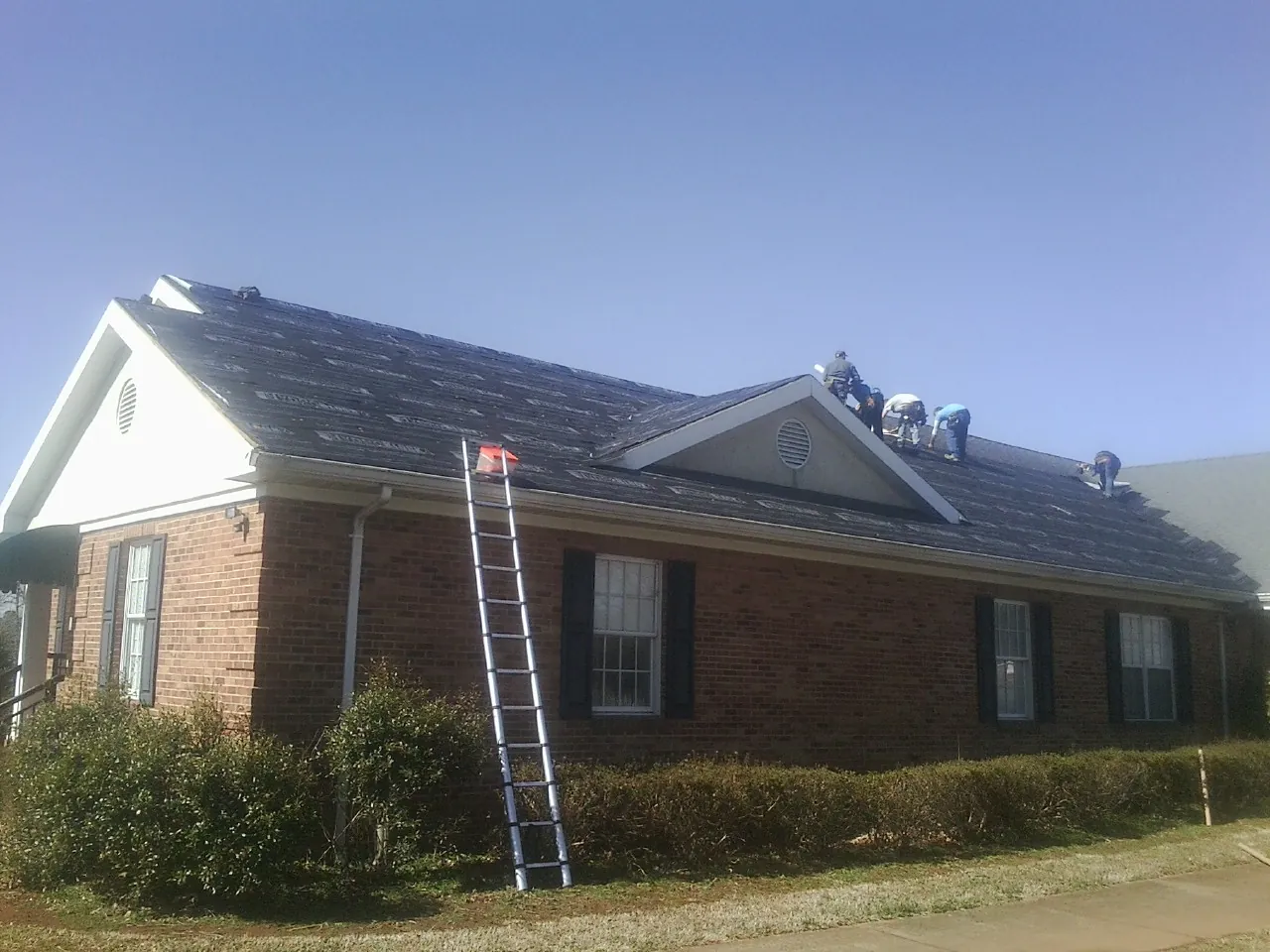 Crew tearing shingles off a brick ranch roof with a ladder set — Metal Roof Repair work by our crew in South Lyon