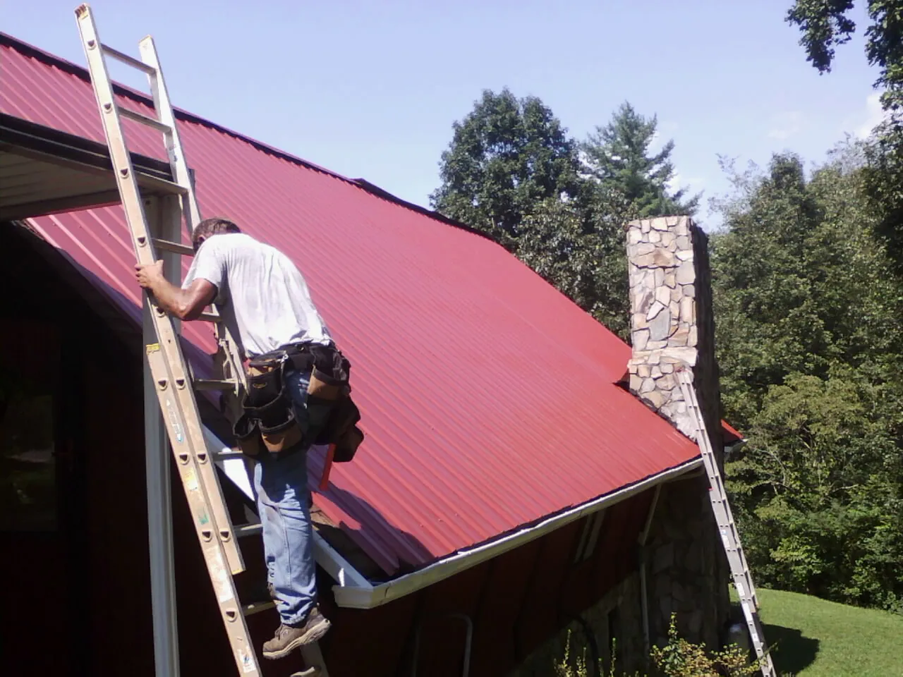 Roofer on a ladder beside a red metal roof and stone chimney — Soffit & Fascia Repair by Roofing Brothers in South Lyon
